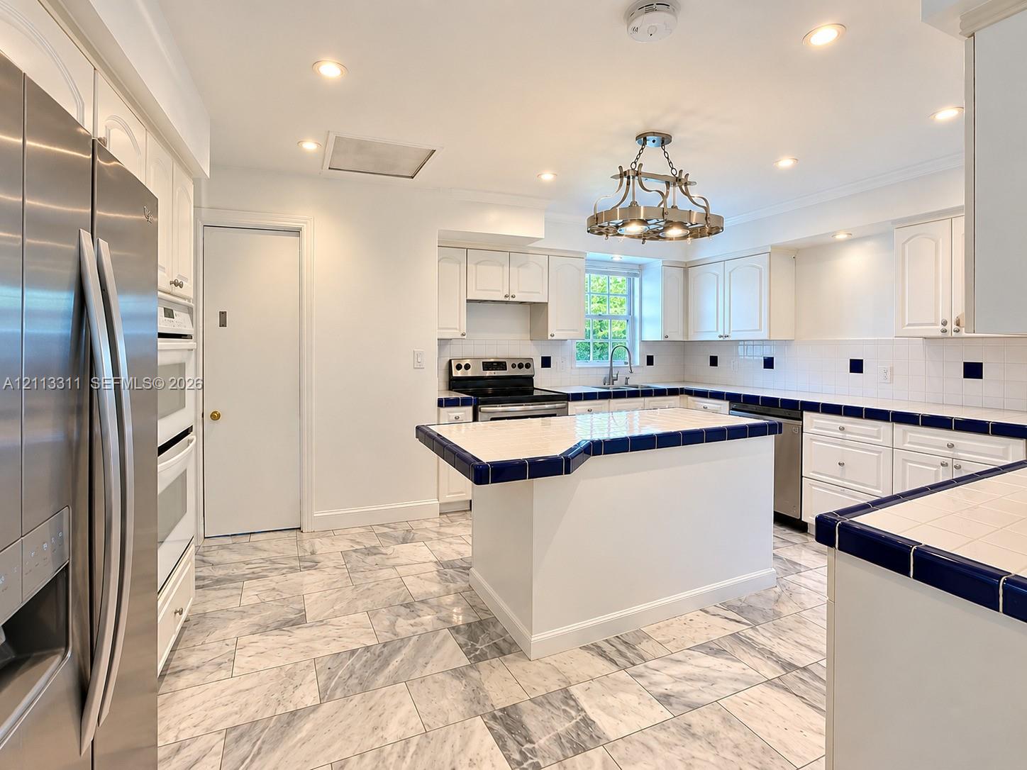 Kitchen featuring stone-tile flooring, white cabinetry, a center island with tiled countertops, stainless steel appliances, and a decorative overhead light fixture