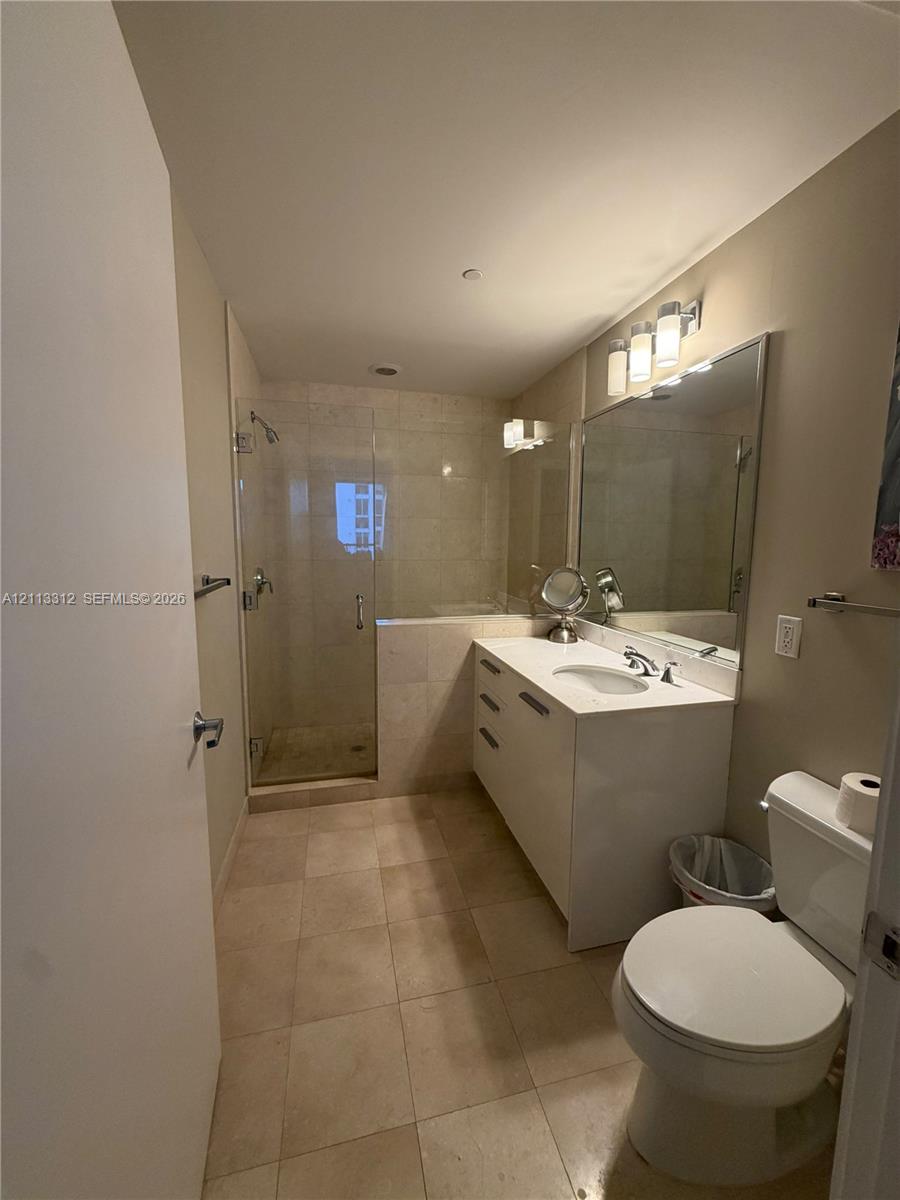 Bathroom featuring a white vanity with an integrated basin, a large wall-mounted mirror, neutral tile flooring, and a glass-enclosed walk-in shower with tile surround