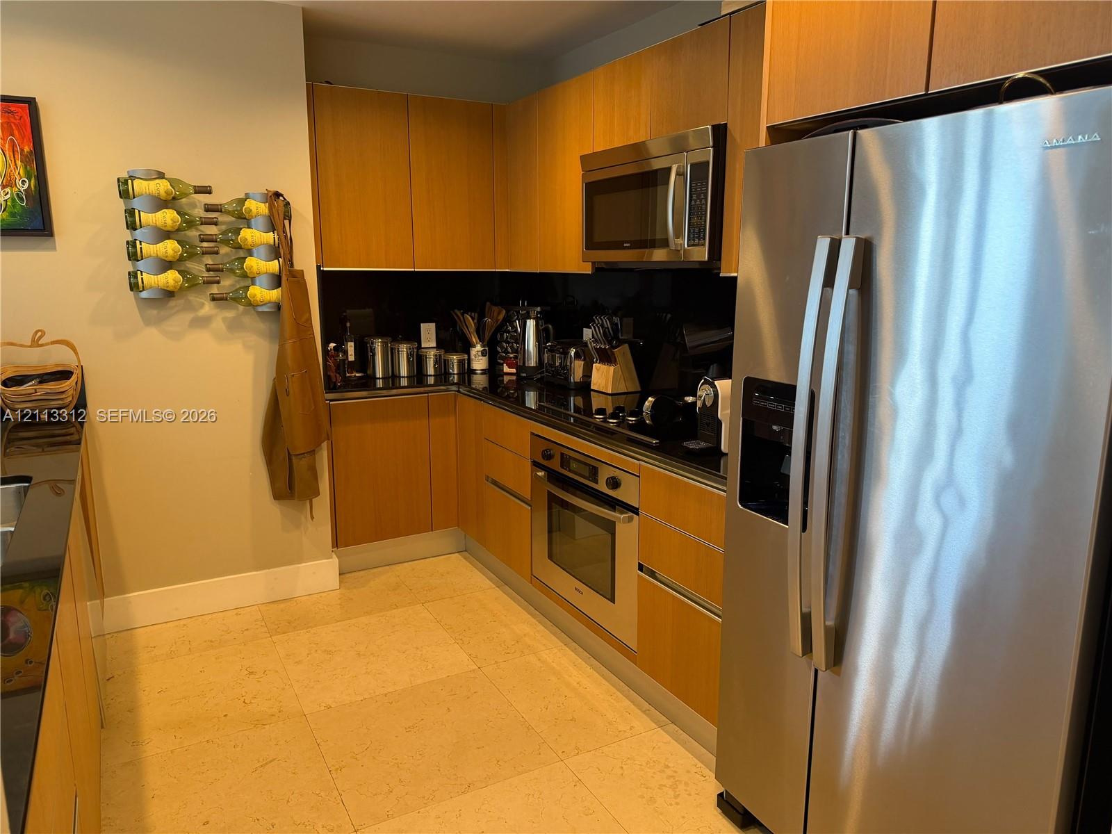 Kitchen featuring wood-veneer flat-panel cabinetry, black countertops, stainless steel wall-mounted oven and microwave, and light-toned stone-finish tile flooring Kitchen featuring wood-veneer flat-panel cabinetry, black countertops, stainless steel wall-mounted oven and microwave, and light-toned stone-finish tile flooring