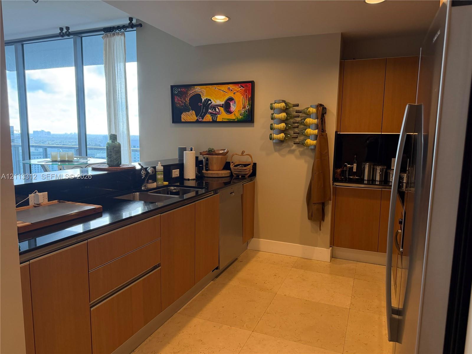 Kitchen featuring flat-panel wood cabinetry, black solid-surface countertops, recessed ceiling lighting, full-height windows, and light-toned stone flooring Kitchen featuring flat-panel wood cabinetry, black solid-surface countertops, recessed ceiling lighting, full-height windows, and light-toned stone flooring
