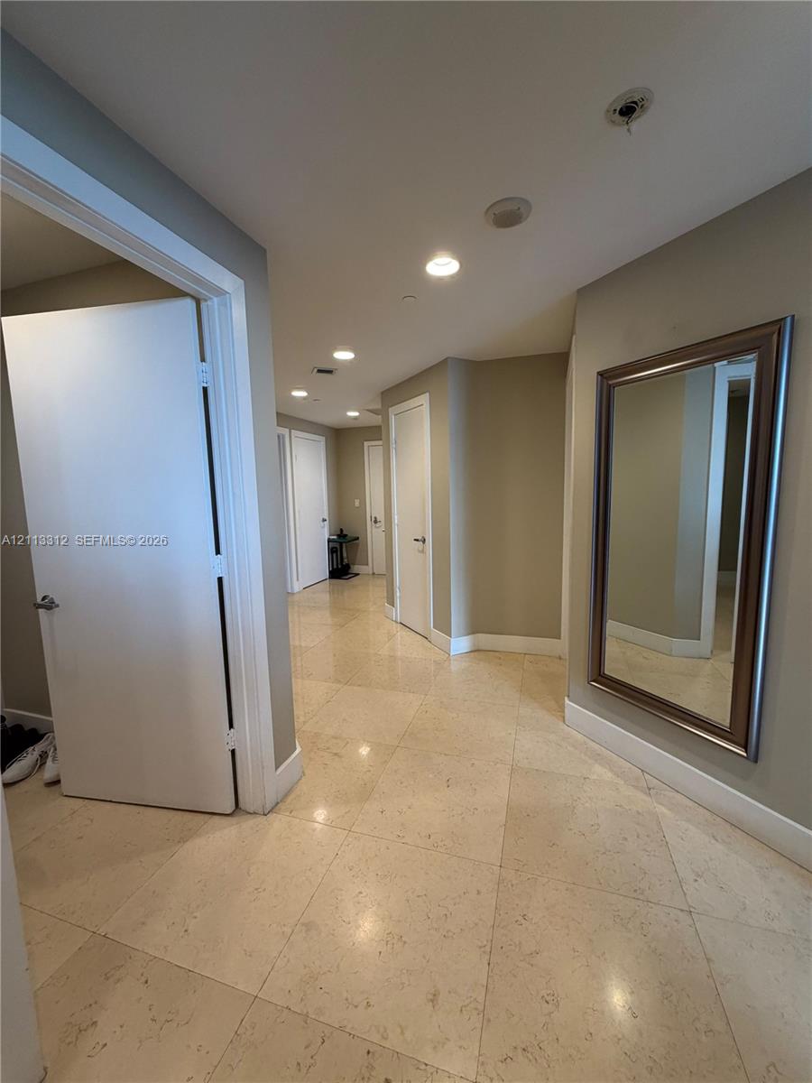 Interior hallway featuring large-format stone tile flooring, recessed ceiling lighting, and solid-core interior doors with metallic hardware Interior hallway featuring large-format stone tile flooring, recessed ceiling lighting, and solid-core interior doors with metallic hardware