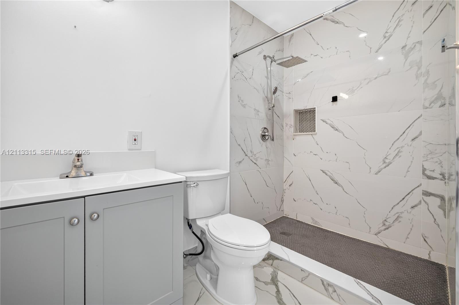 Bathroom featuring a grey vanity with a white integrated sink, walk-in shower with floor-to-ceiling stone-look tile, rain-style shower head, and built-in recessed wall niche