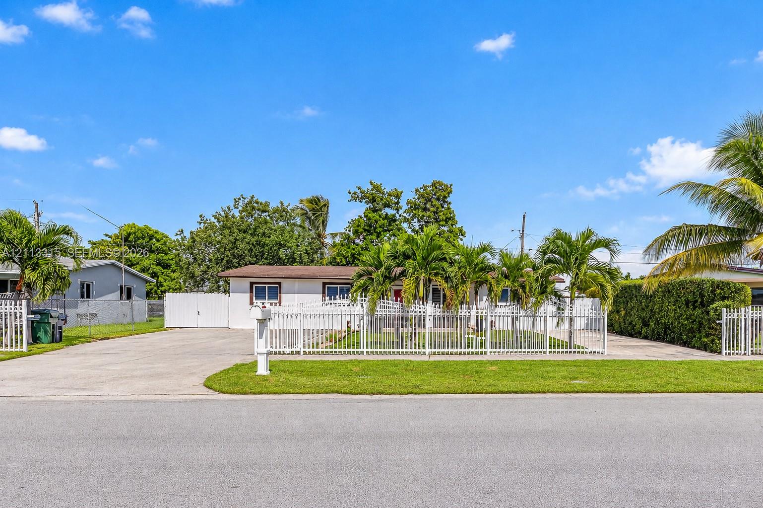 Single-story residence featuring a white exterior, expansive concrete driveway, full-property white metal fencing, and mature palm trees