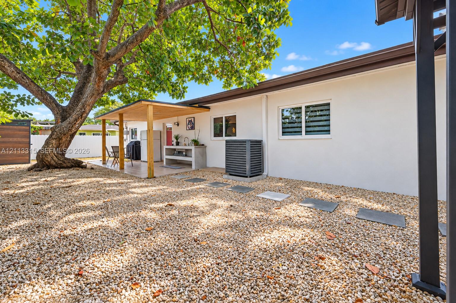 Backyard featuring a gravel-surfaced ground, a covered patio with a wooden support structure, a built-in outdoor counter with a sink, and mature shade trees