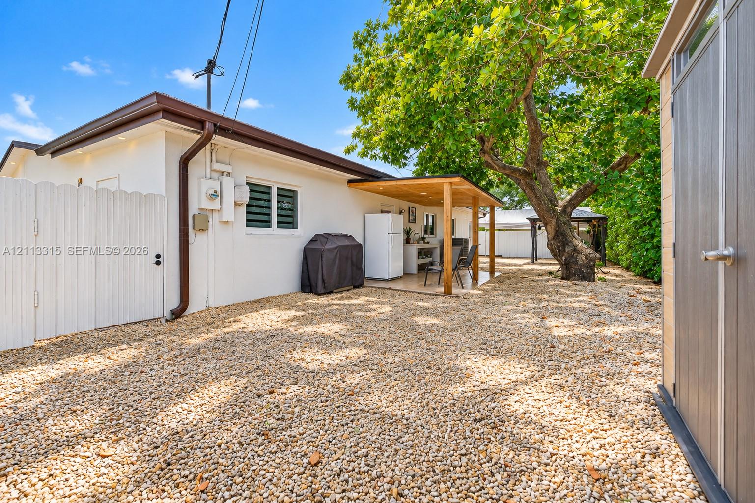 Low-maintenance gravel yard featuring a covered patio, built-in outdoor counter, white privacy fencing, and mature trees