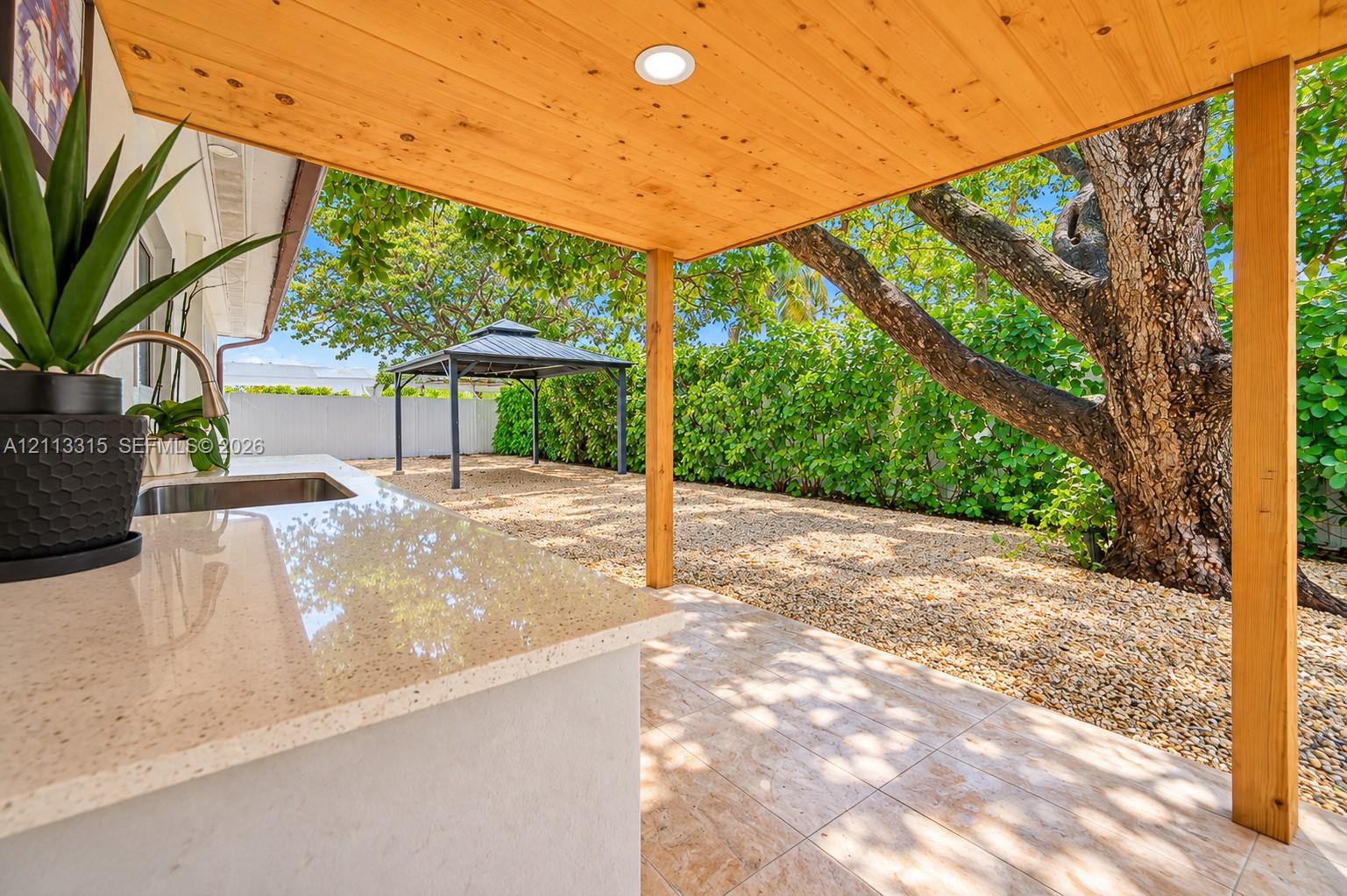 Covered outdoor kitchen featuring a light-toned solid surface countertop and undermount sink, complemented by a wood-finished ceiling, recessed lighting, and tiled patio flooring