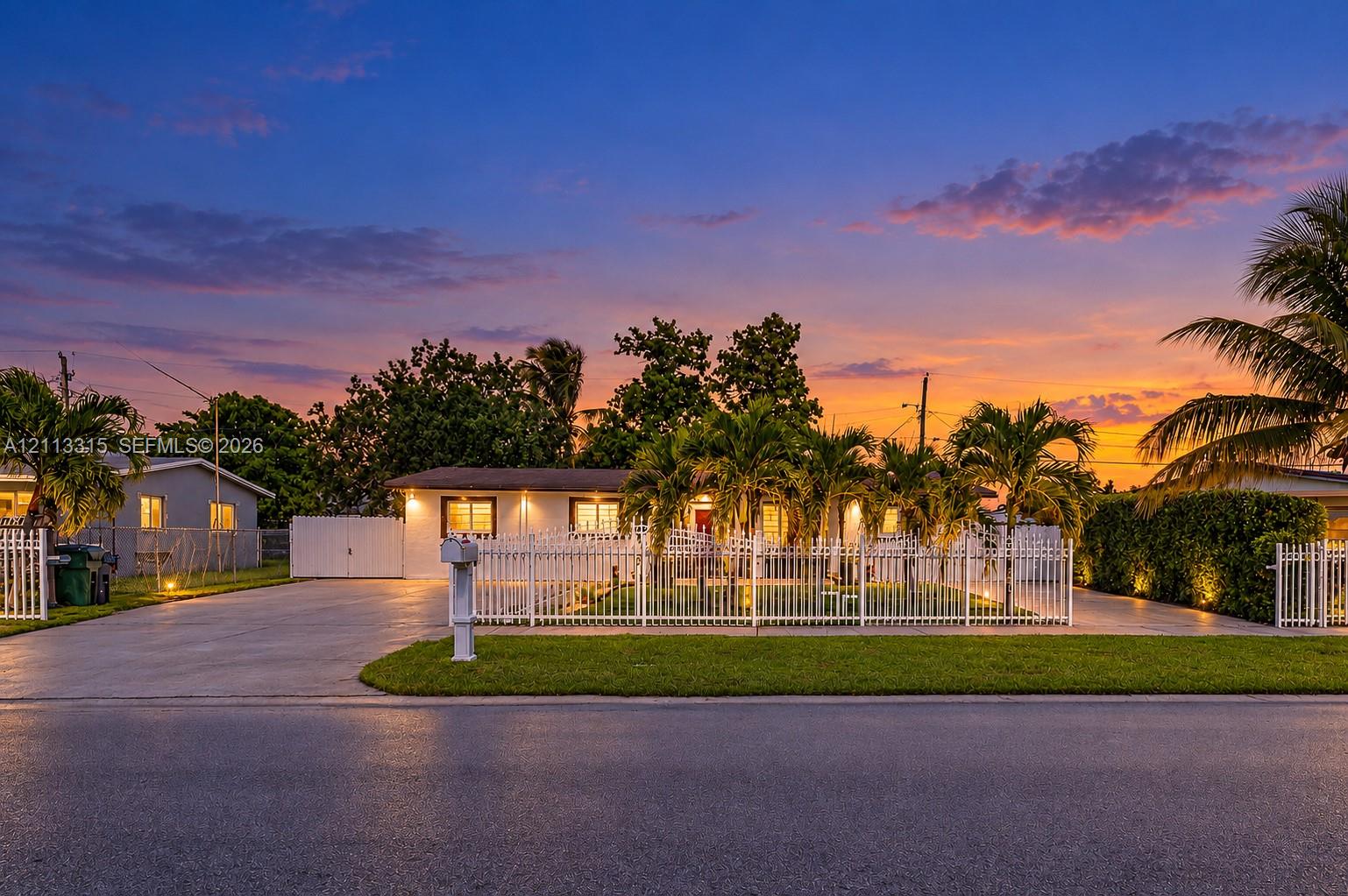 Single-story residence featuring a painted masonry exterior, expansive concrete driveway, full-perimeter white metal fencing, and mature palm landscaping Single-story residence featuring a painted masonry exterior, expansive concrete driveway, full-perimeter white metal fencing, and mature palm landscaping