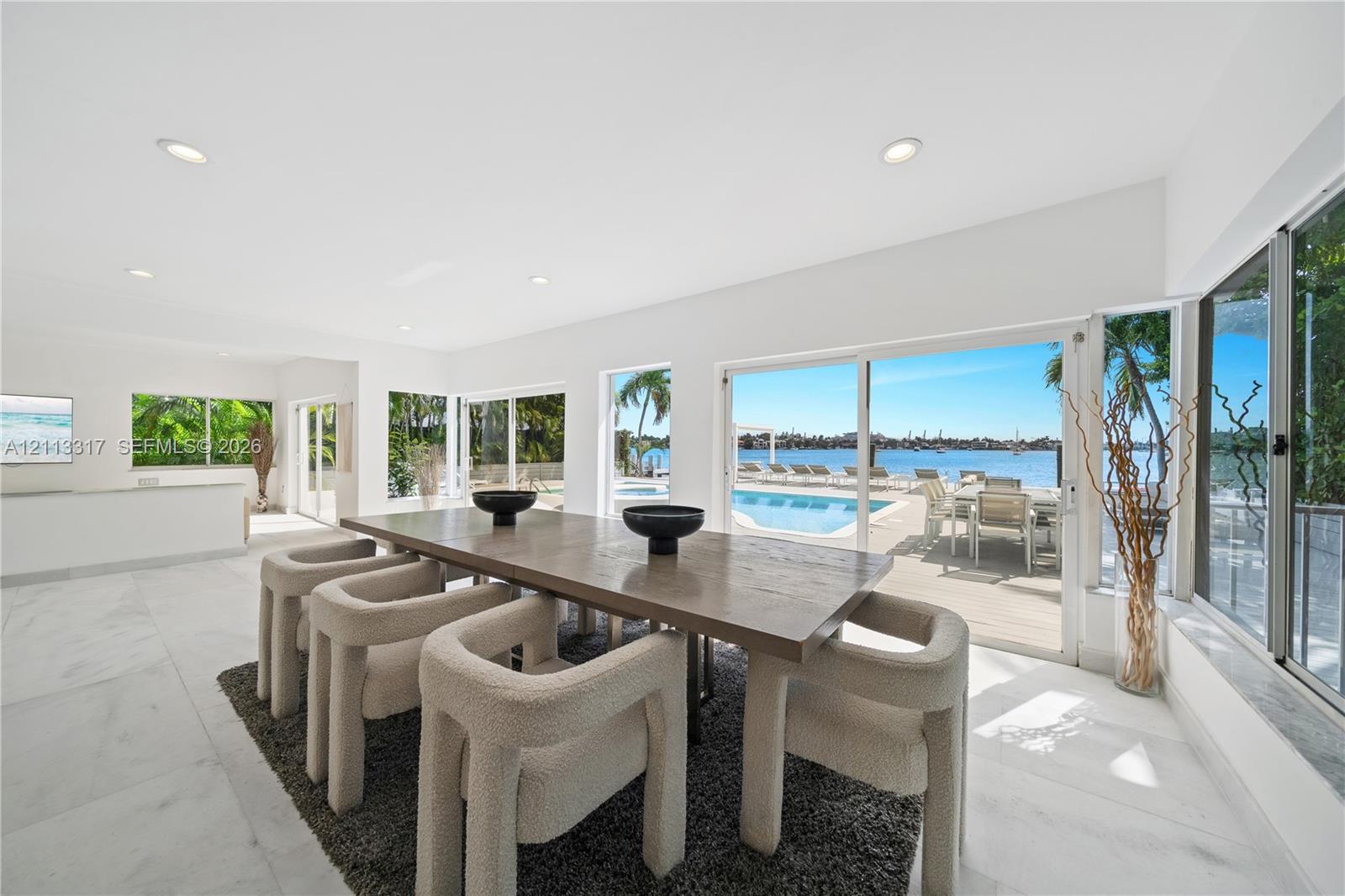 Open-concept interior featuring large-format tile flooring, recessed lighting, and expansive floor-to-ceiling glass sliding doors opening to an outdoor pool and waterfront Open-concept interior featuring large-format tile flooring, recessed lighting, and expansive floor-to-ceiling glass sliding doors opening to an outdoor pool and waterfront