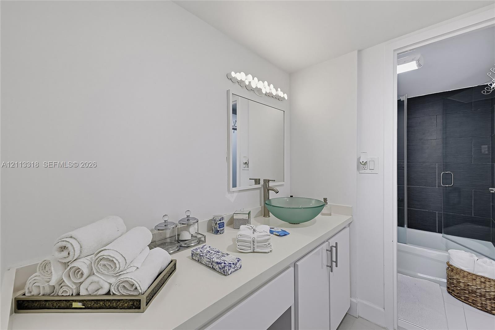 Bathroom featuring a solid-surface countertop, glass vessel sink with brushed-finish faucet, white cabinetry with silver-tone hardware, and a tub-shower combination with dark wall tile