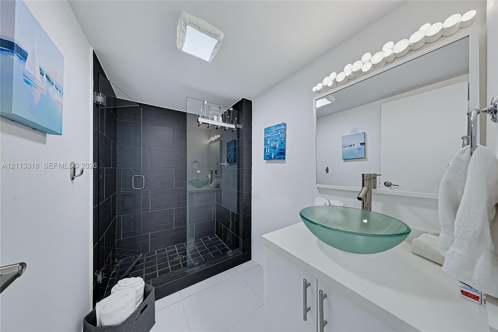 Walk-in shower with dark tile surround and glass enclosure, glass vessel sink, white vanity, and multi-bulb wall-mounted lighting