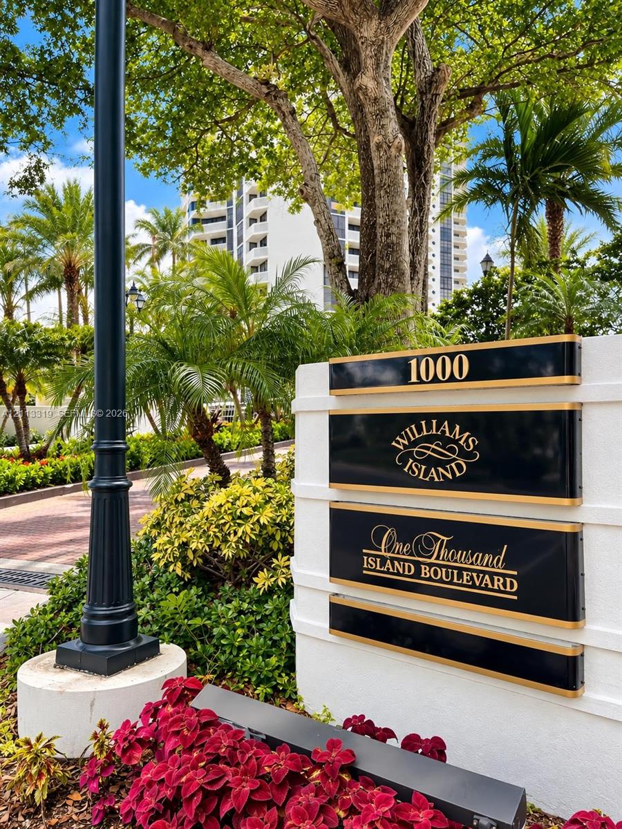 Monument signage featuring gold-lettered panels on a white stucco base, surrounded by mature palm trees, professional landscaping, and a brick-paved entry drive