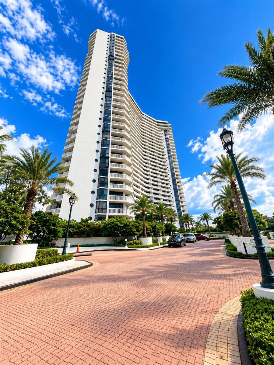 High-rise building featuring a white facade, curvilinear architectural design, stacked private balconies, and a paved brick entrance drive lined with mature palm trees and decorative lighting fixtures