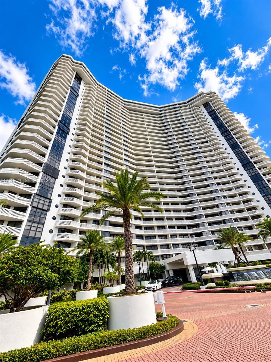 High-rise architectural exterior featuring tiered private balconies, floor-to-ceiling glass paneling, a porte-cochere entryway, and a landscaped circular motor court with paver surfacing
