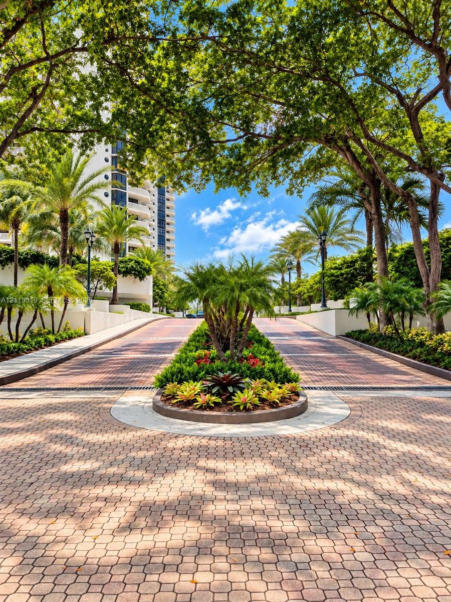 Paved circular drive featuring a central garden bed, mature canopy trees, palm landscaping, and decorative brick-style pavers
