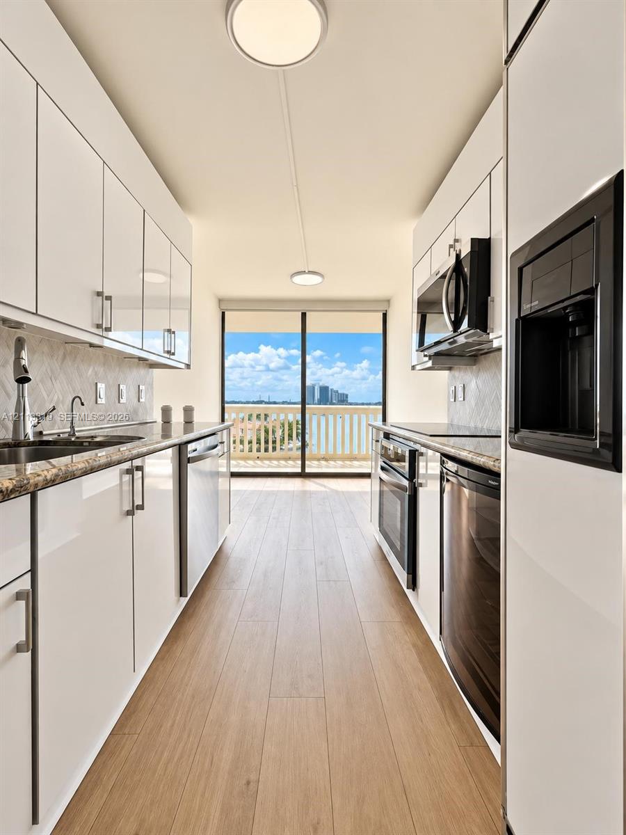 Galley-style kitchen featuring high-gloss white cabinetry, stone-surface countertops, wood-finish flooring, built-in stainless steel appliances, and glass sliding doors providing exterior access