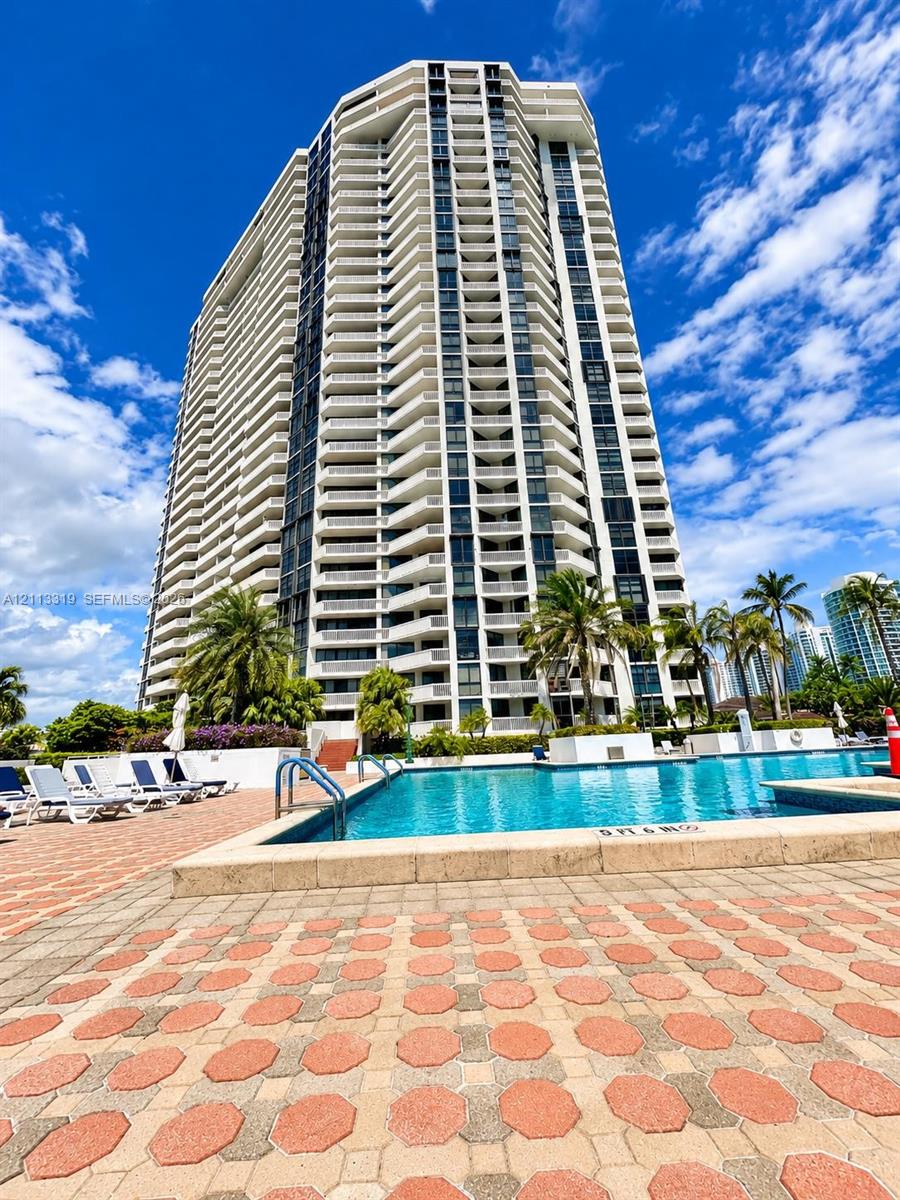 High-rise building featuring stacked balconies and vertical glass elements, with an outdoor swimming pool set in a paved stone deck surrounded by palm trees