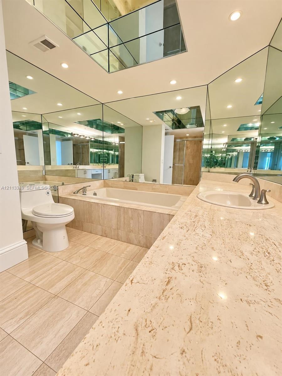 Stone-surfaced vanity with an integrated basin, inset soaking tub, tile flooring, and wall-to-ceiling mirror panels