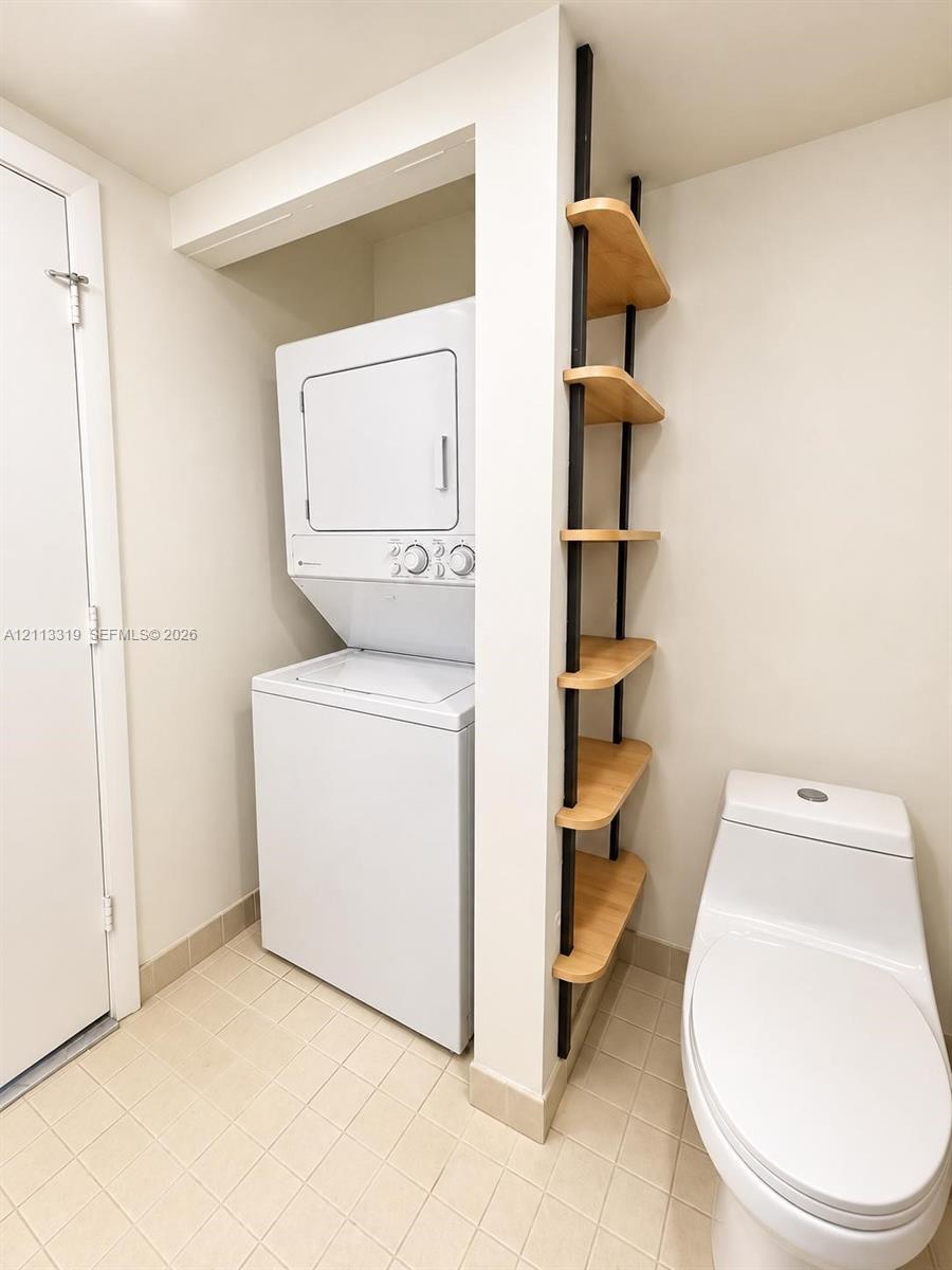 Stacked laundry unit with wall-mounted shelving featuring natural wood-finish planks and metal supports, paired with a white porcelain toilet and tile flooring