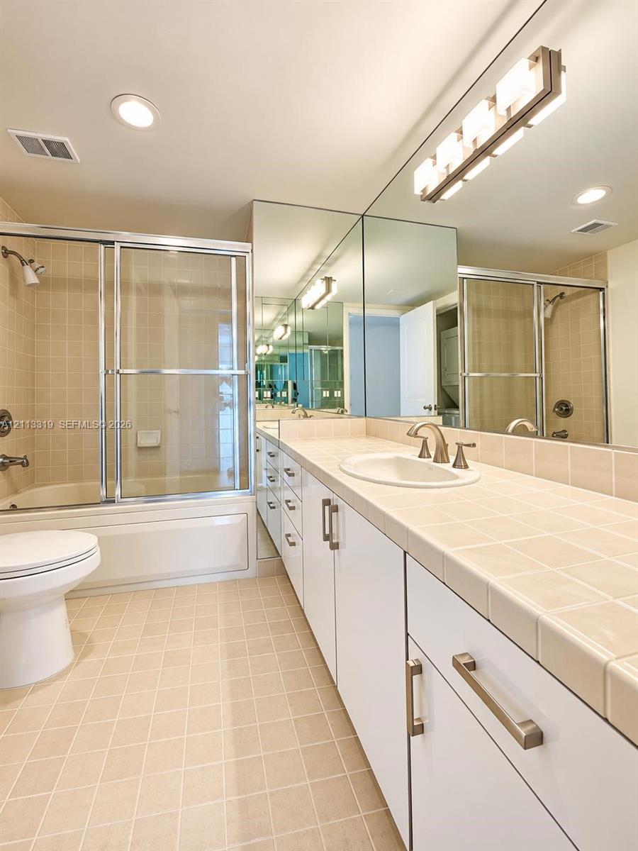 Bathroom featuring a white vanity cabinet with tile countertop, integrated sink, full-width wall mirror, and tub-shower combination with sliding glass doors