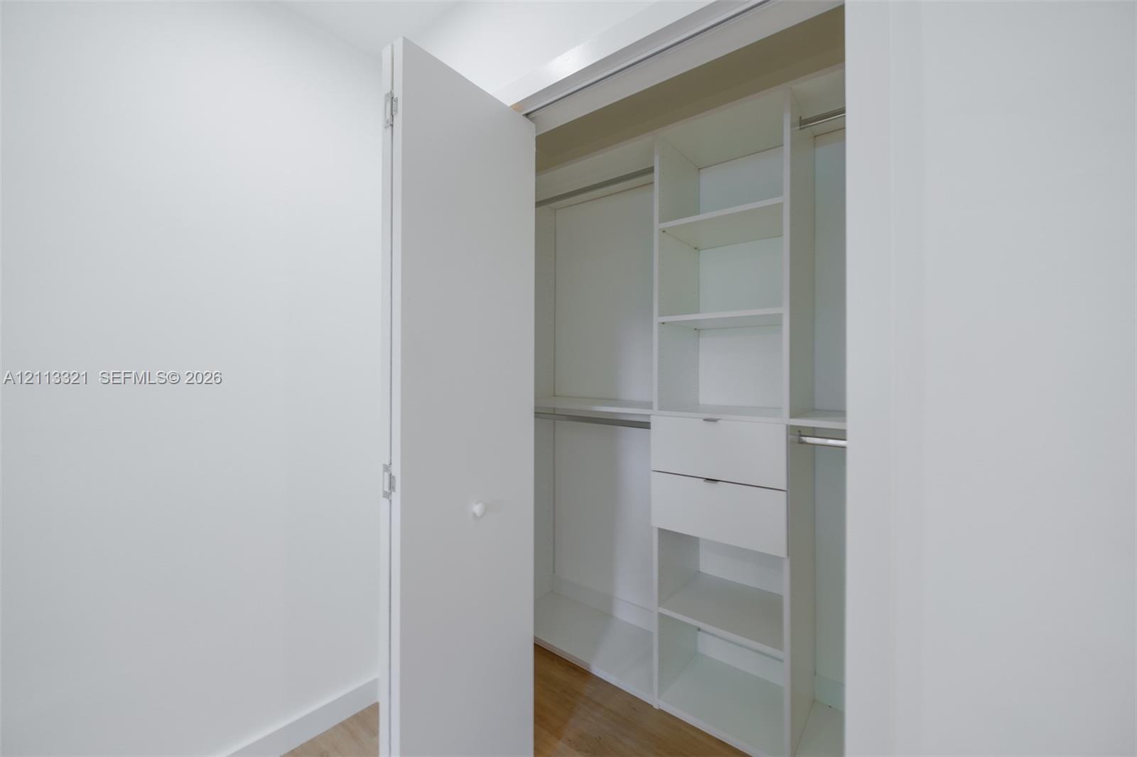 Custom closet featuring white built-in cabinetry, dual drawers, integrated shelving, and metal hanging rods over wood-finish flooring