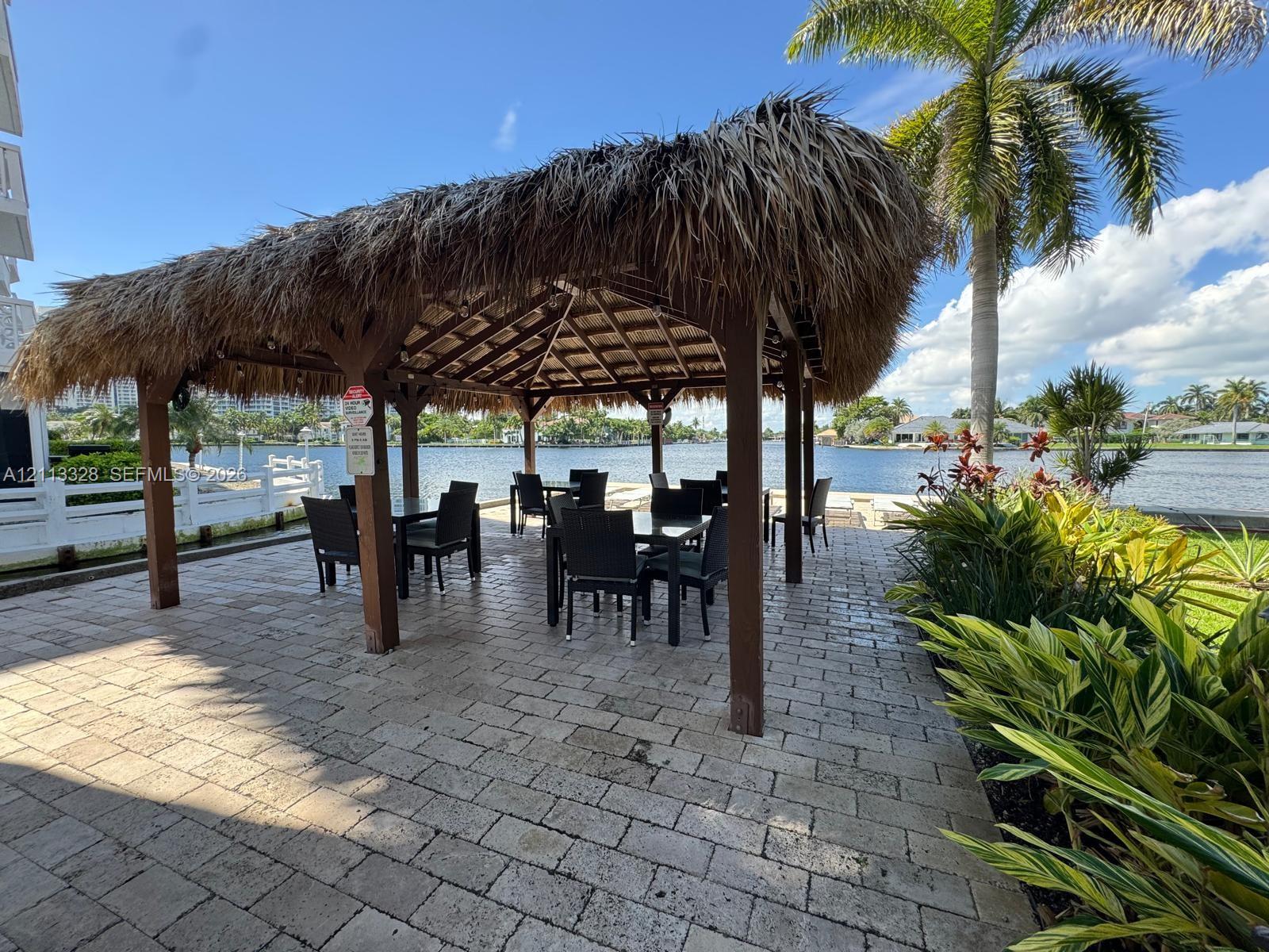 Waterfront gazebo with a thatched roof, supported by timber posts, situated on a stone paver patio