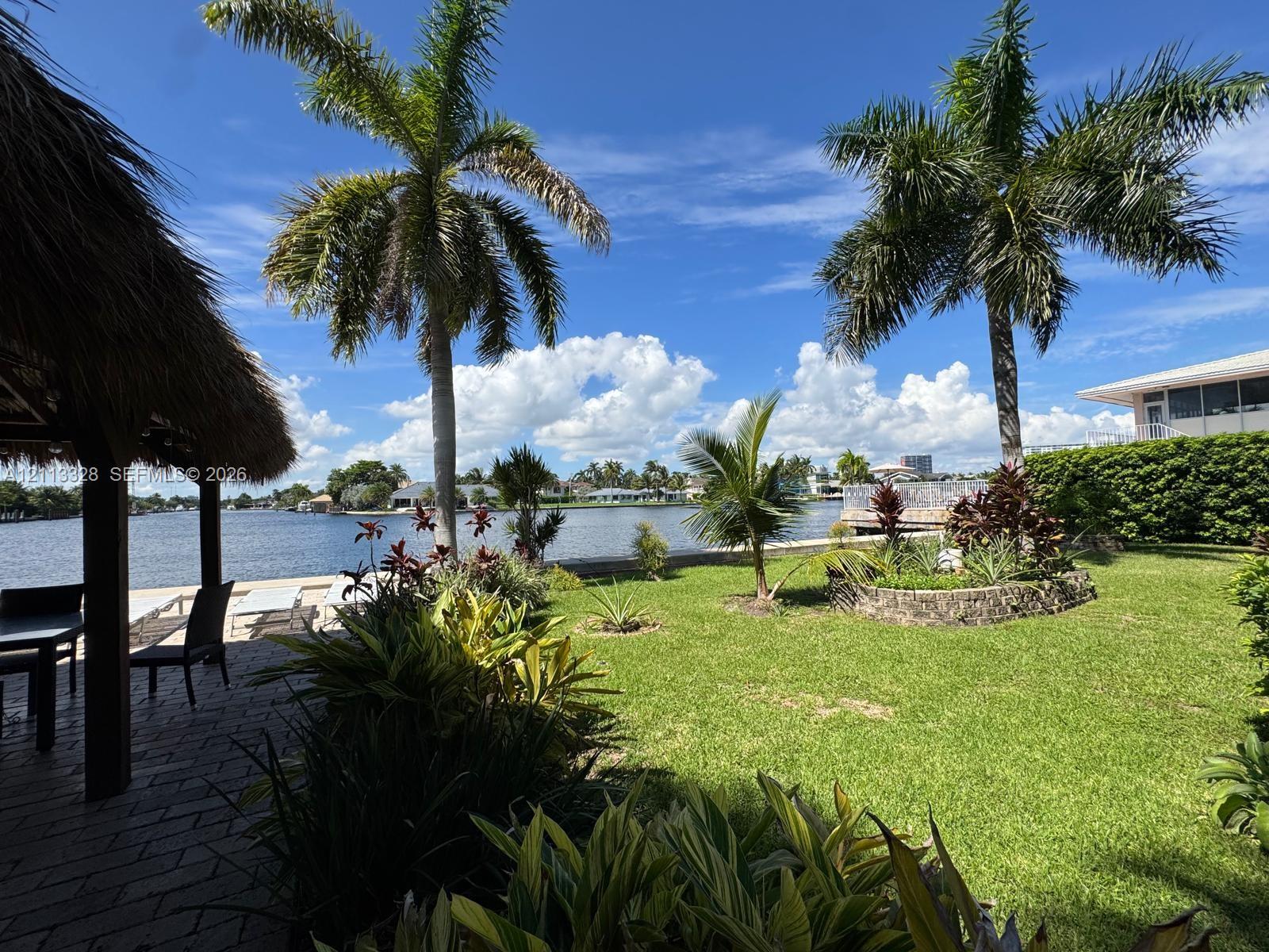 Waterfront backyard featuring a thatched-roof tiki hut, paved patio, expansive lawn, mature palm trees, and a concrete seawall Waterfront backyard featuring a thatched-roof tiki hut, paved patio, expansive lawn, mature palm trees, and a concrete seawall