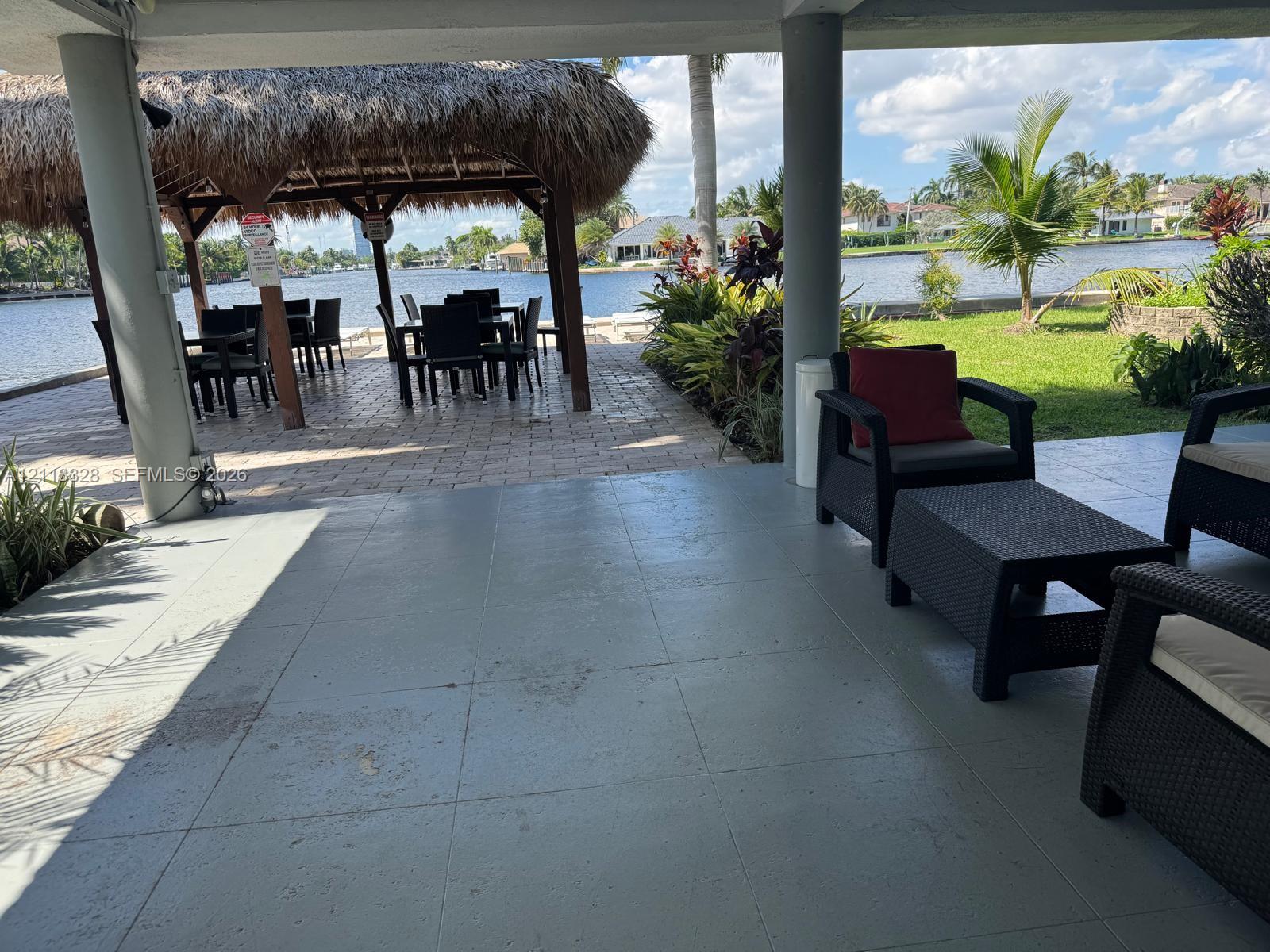 Covered patio with tile flooring and structural columns, featuring a waterfront paver area, a thatched-roof tiki hut, and mature landscaping