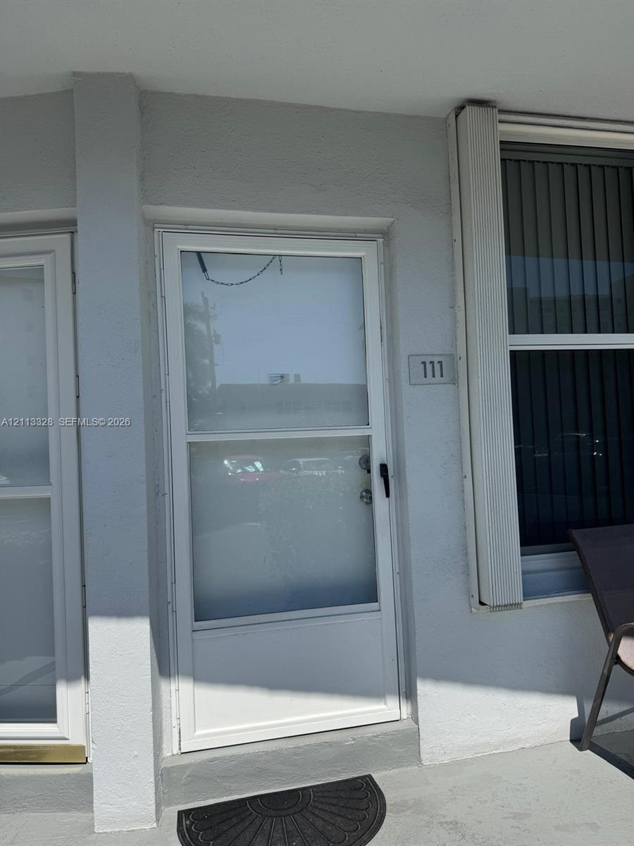 Exterior entry featuring a glass-panel storm door, light-colored smooth stucco siding, vertical window casing, and a dedicated address plaque