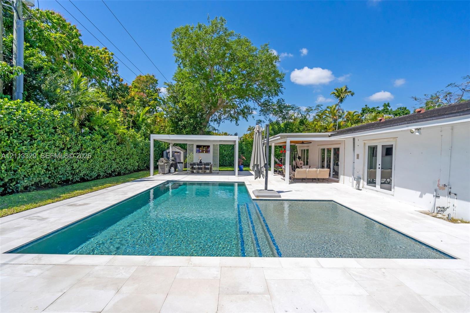 Swimming pool with a sun shelf and built-in steps, surrounded by expansive light-toned stone patio pavers, featuring a detached covered pavilion and a covered exterior porch