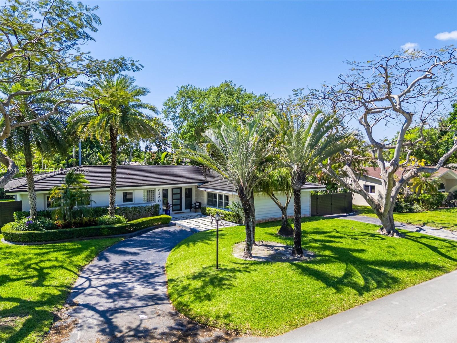 Single-story residence featuring a dark-tiled hip roof, paved circular driveway, manicured landscaping, and mature palm trees