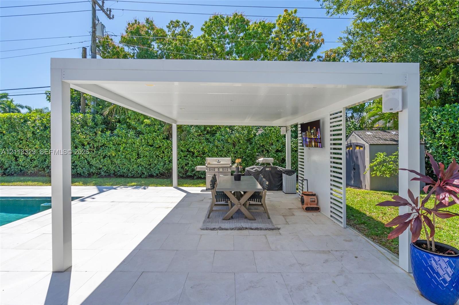 Covered outdoor living space featuring a white aluminum pergola, light-toned stone patio flooring, built-in recessed lighting, and a slatted privacy screen