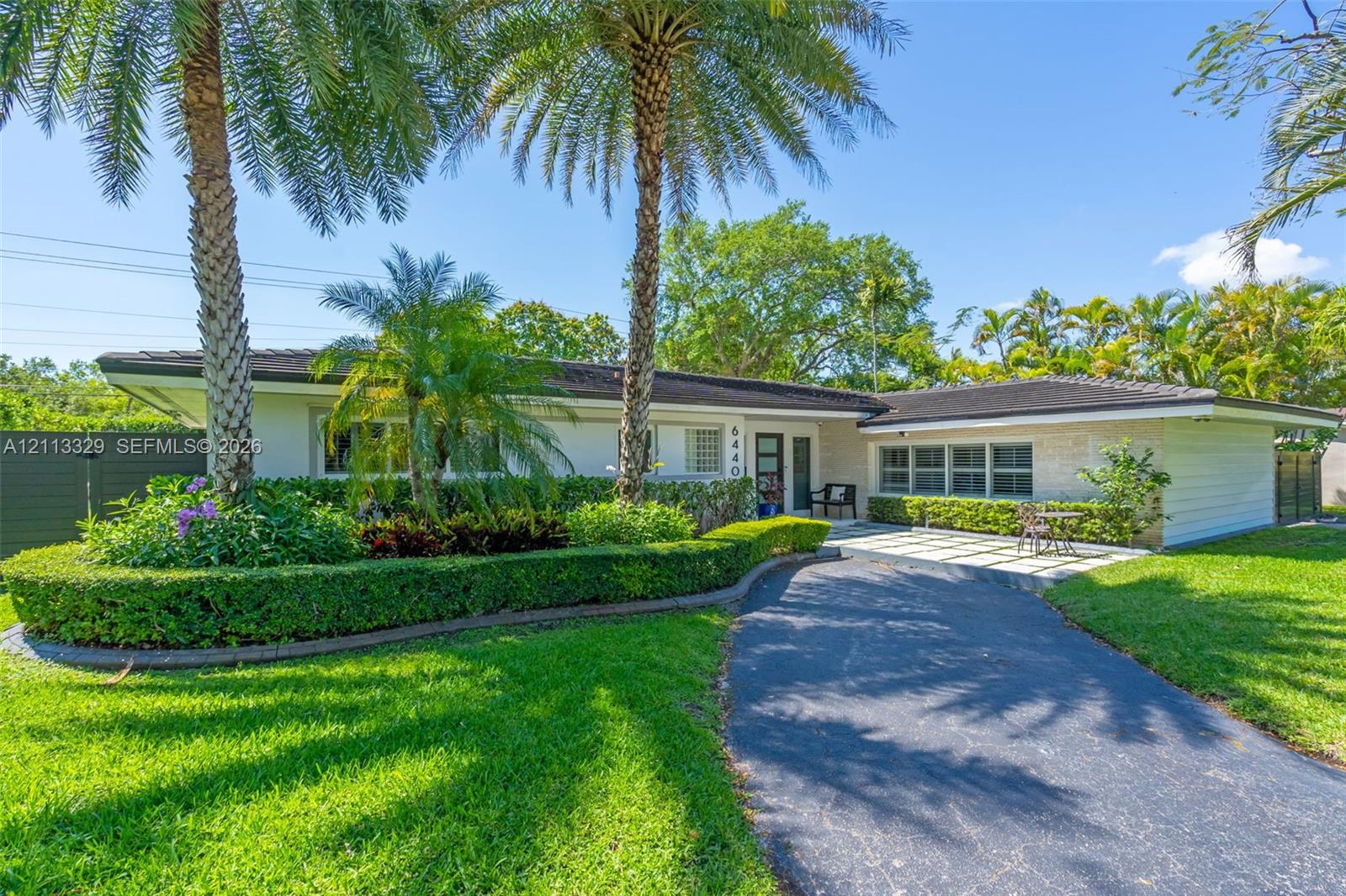 Single-story residence featuring a low-pitched roof, manicured hedges, mature palm trees, stone-finish exterior accents, and a paved front patio area