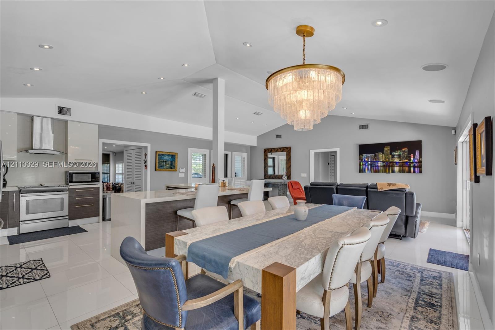 Vaulted ceilings with recessed lighting, large central kitchen island with waterfall countertop, high-gloss cabinetry, stainless steel range with overhead ventilation, and a tiered crystal chandelier