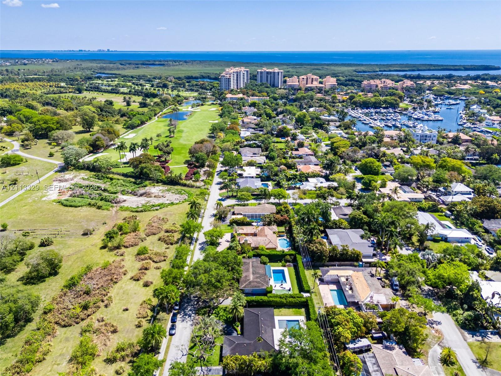 Aerial perspective of a coastal residential area featuring mature landscaping, proximity to a golf course, expansive open green spaces, and a maritime marina