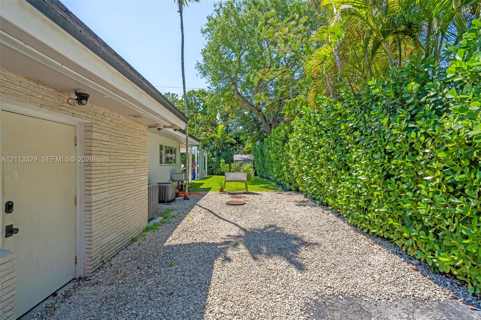 Side exterior featuring painted brick siding, gravel ground cover, and a privacy hedge of mature greenery