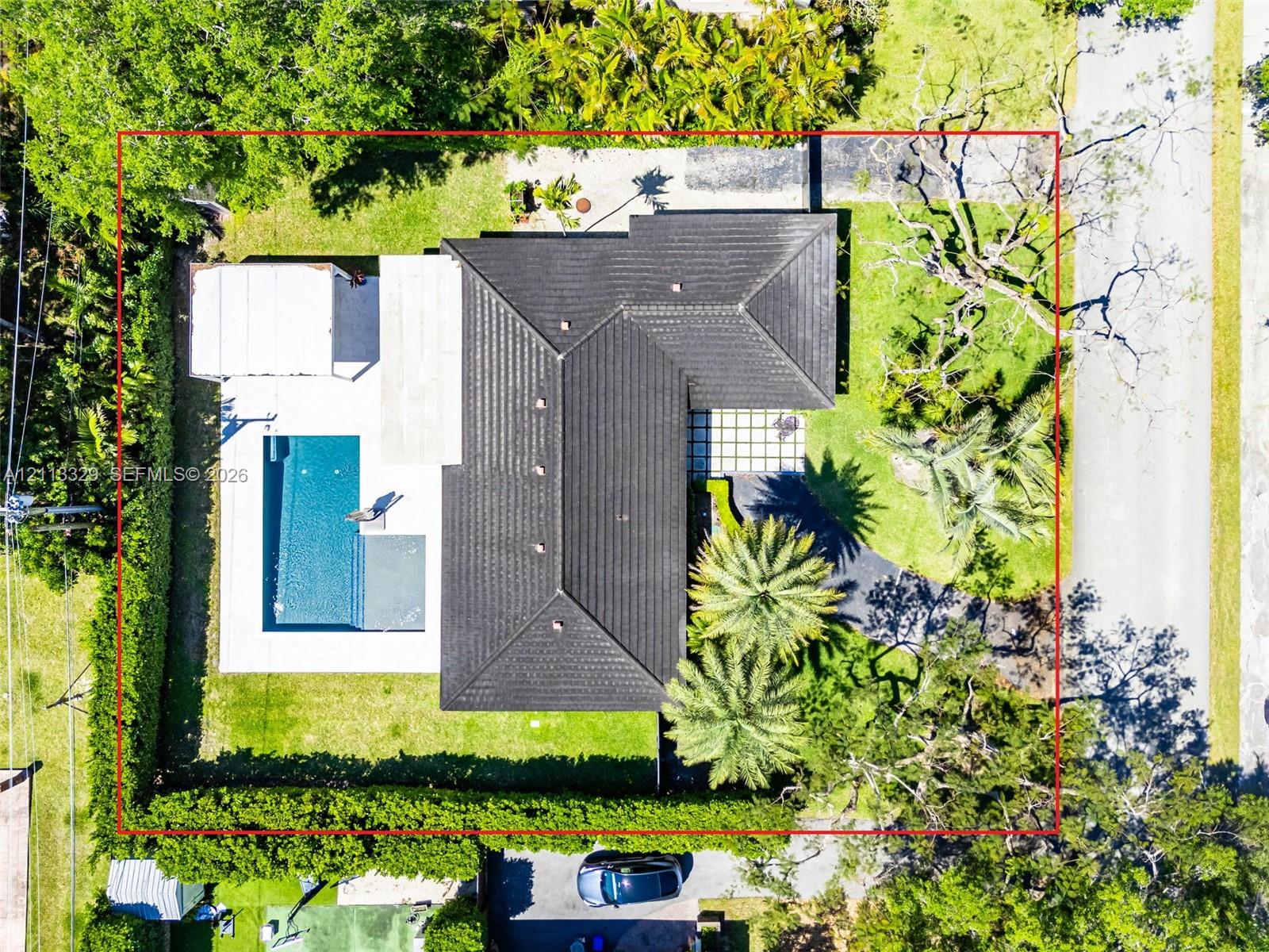 Aerial perspective of a property featuring a tiled hip roof, rectangular swimming pool, concrete patio, asphalt driveway, and mature landscaping