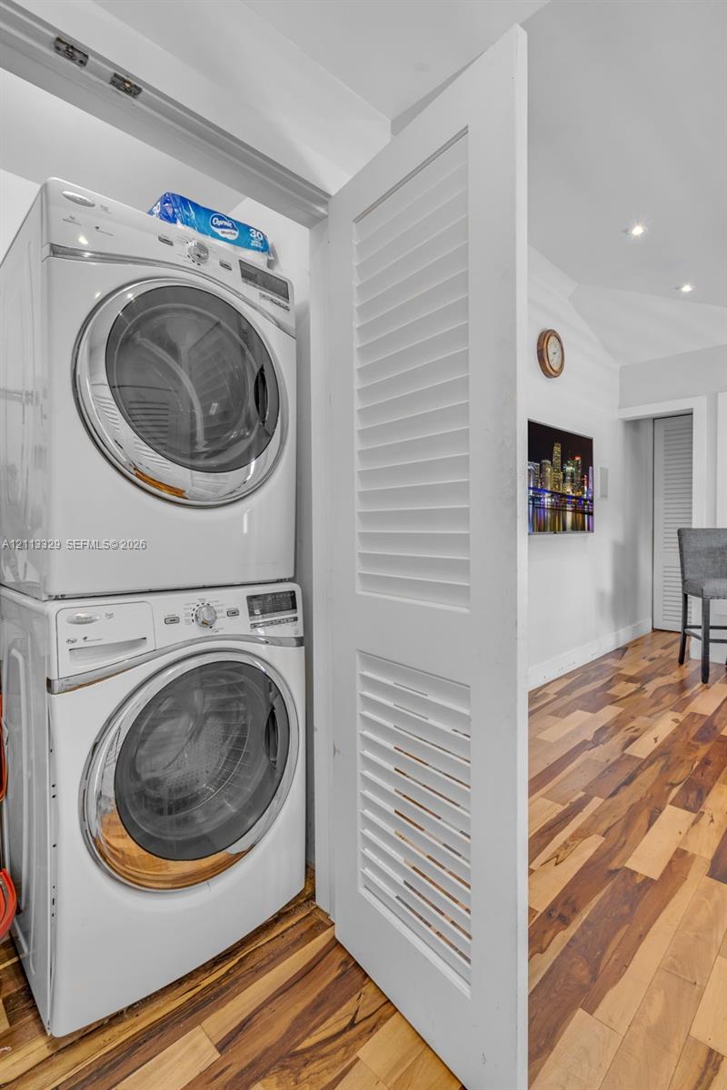 Stacked laundry unit concealed by a white louvered door, complemented by wood-finish flooring and recessed ceiling lighting