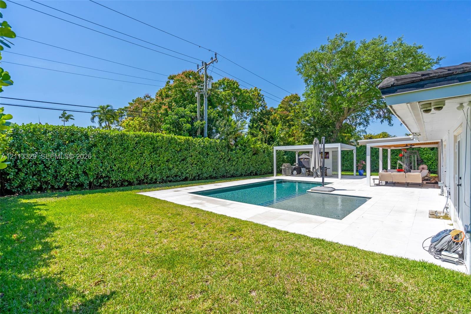 Backyard featuring an inground swimming pool, light-colored stone patio, covered pergola, and privacy hedges