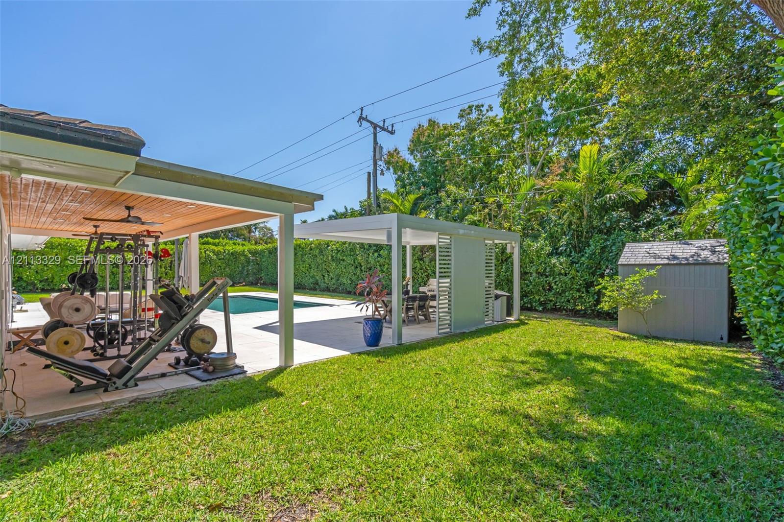 Backyard featuring a swimming pool, a covered patio with a beadboard ceiling, a separate metal-framed pergola, and a storage shed surrounded by mature perimeter hedging