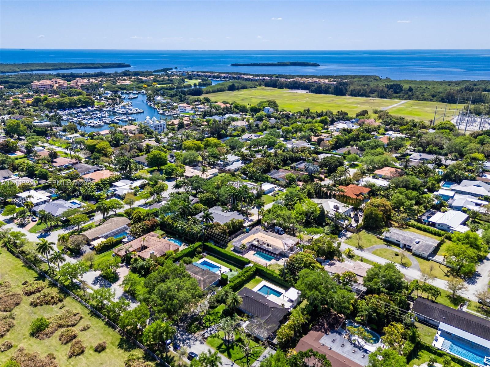 Aerial perspective of a coastal residential neighborhood featuring extensive tree canopy, manicured lawns, private swimming pools, and proximity to a marina and open water
