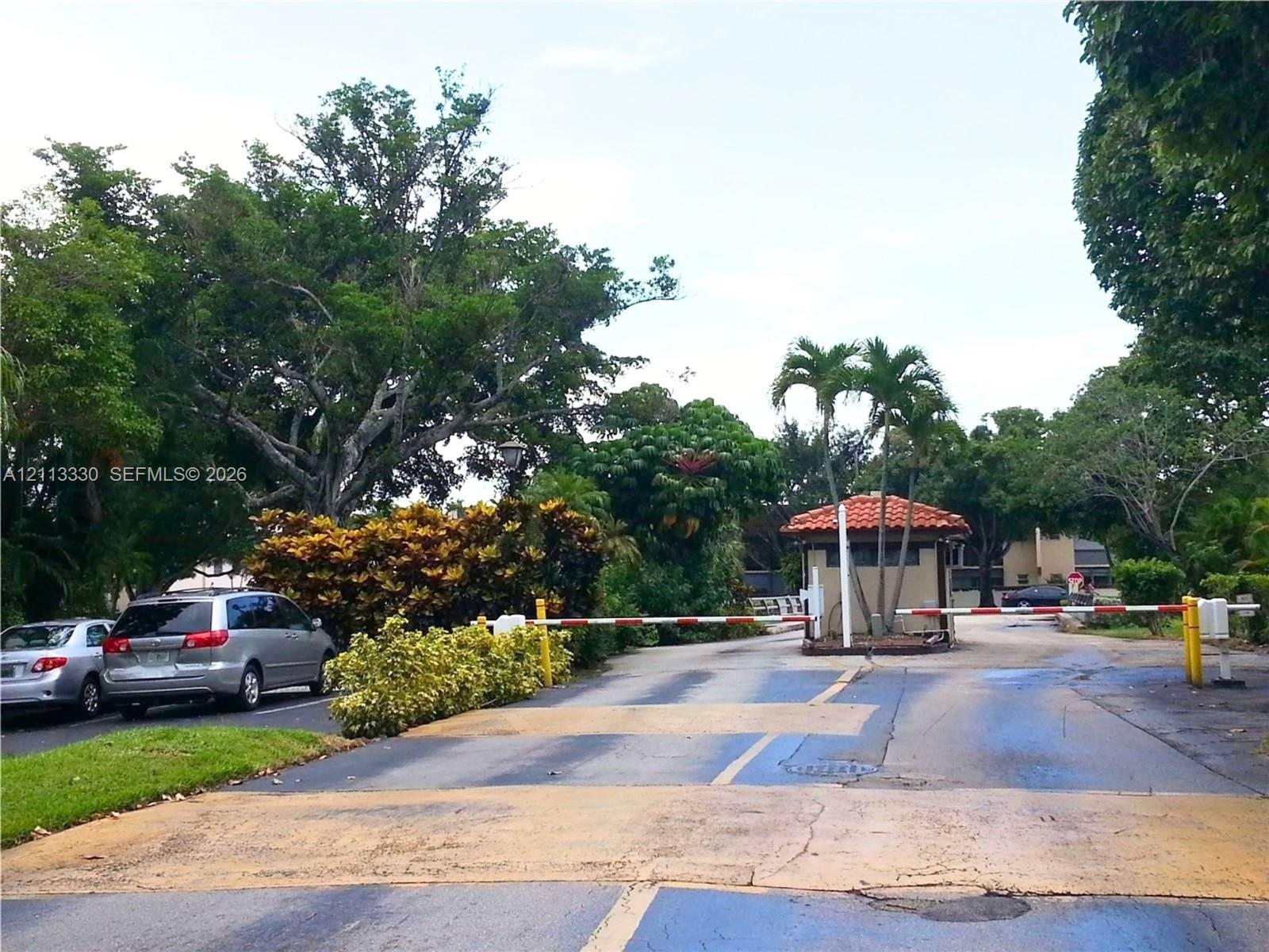 Gated entry featuring a controlled-access barrier arm, guard house with a terracotta tile roof, and mature tropical landscaping Gated entry featuring a controlled-access barrier arm, guard house with a terracotta tile roof, and mature tropical landscaping