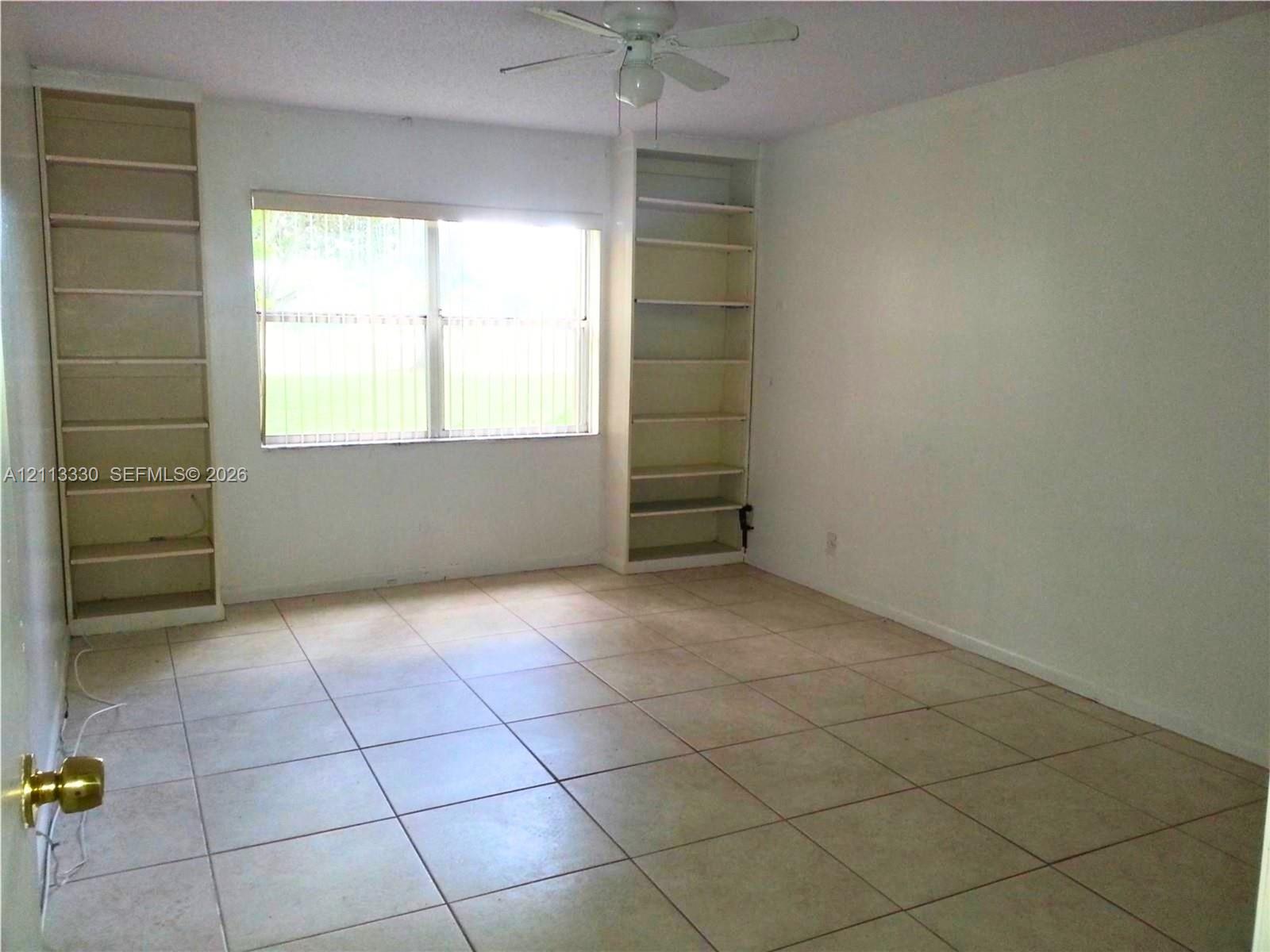 Room featuring neutral tile flooring, dual floor-to-ceiling built-in shelving units, and a ceiling fan Room featuring neutral tile flooring, dual floor-to-ceiling built-in shelving units, and a ceiling fan