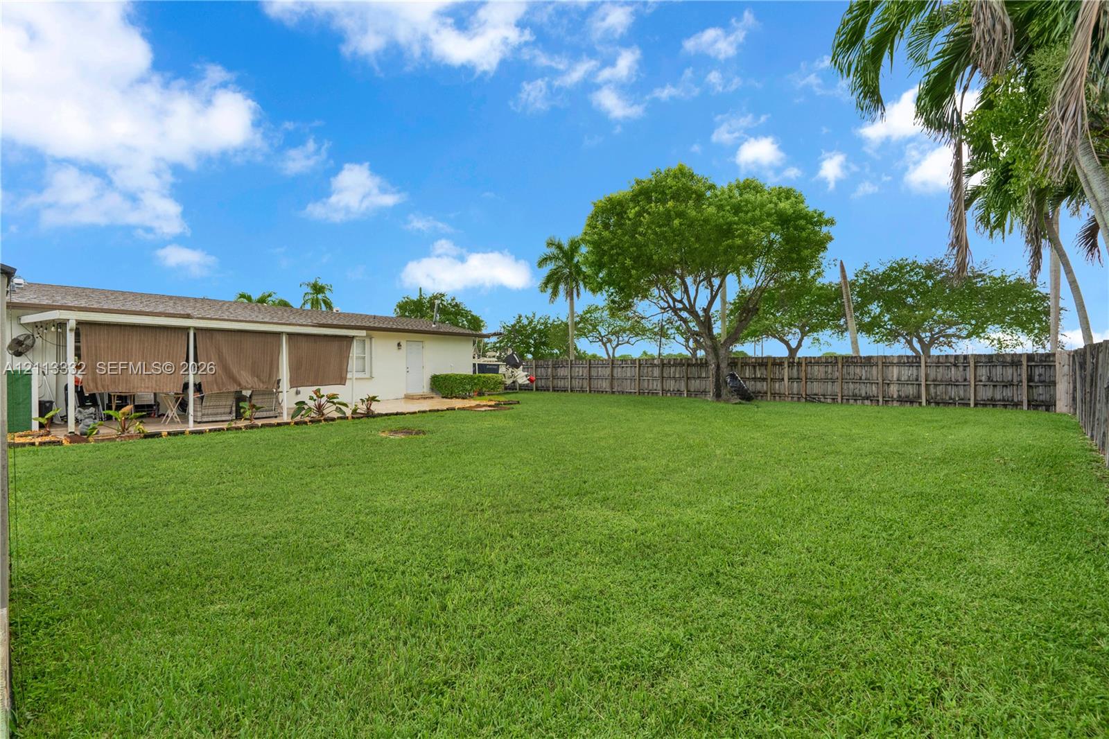 Expansive backyard featuring a large grass lawn, perimeter wood fencing, mature trees, and a covered patio area