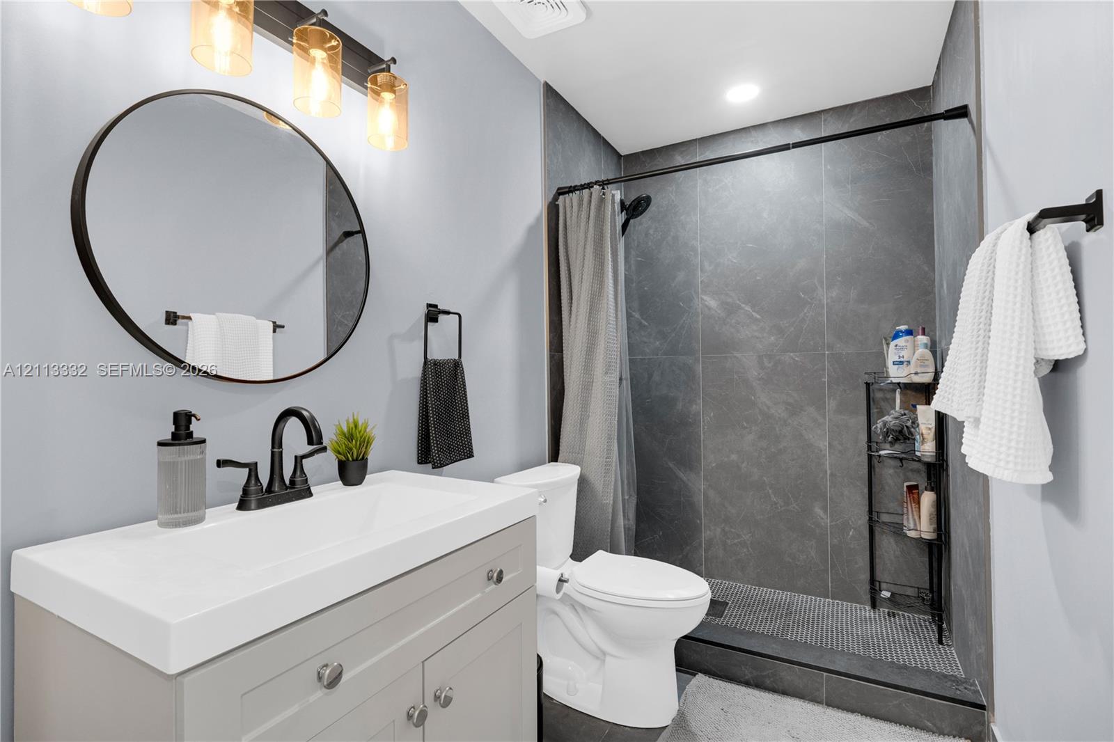 Bathroom featuring a vanity with a rectangular sink, matte black hardware, a round mirror, and a walk-in shower with dark tile surround