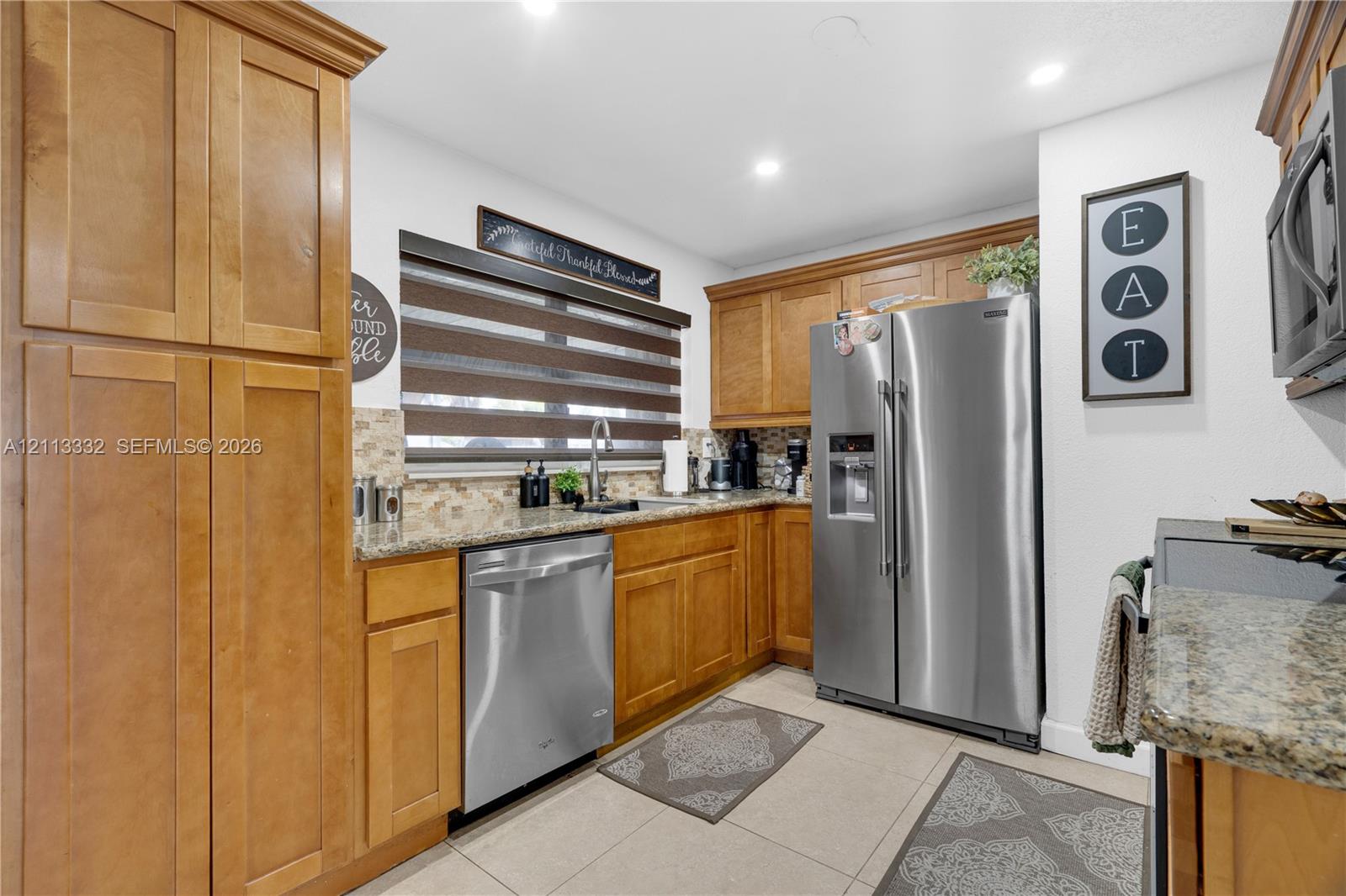 Kitchen featuring medium-toned wood cabinetry, stone countertops, tiled backsplash, stainless steel appliances, and large-format floor tile