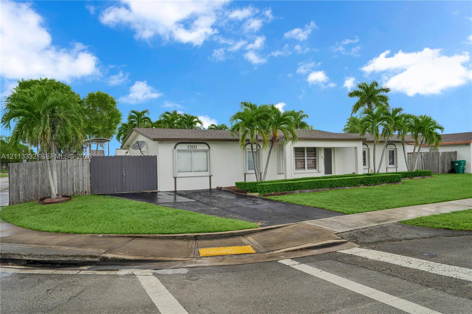 Single-story exterior featuring a light-toned stucco facade, paved driveway, mature palm trees, and perimeter privacy fencing