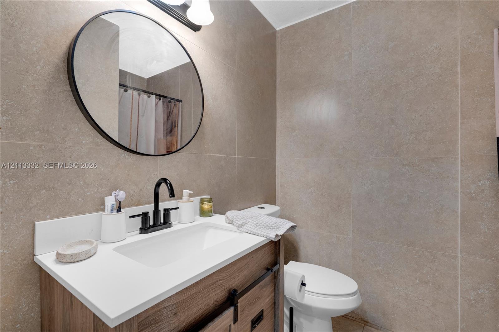 Bathroom featuring a vanity with a white countertop and wood-finish cabinetry, an oval mirror with a matte black frame, a matte black faucet, and full-height stone-finish wall tile