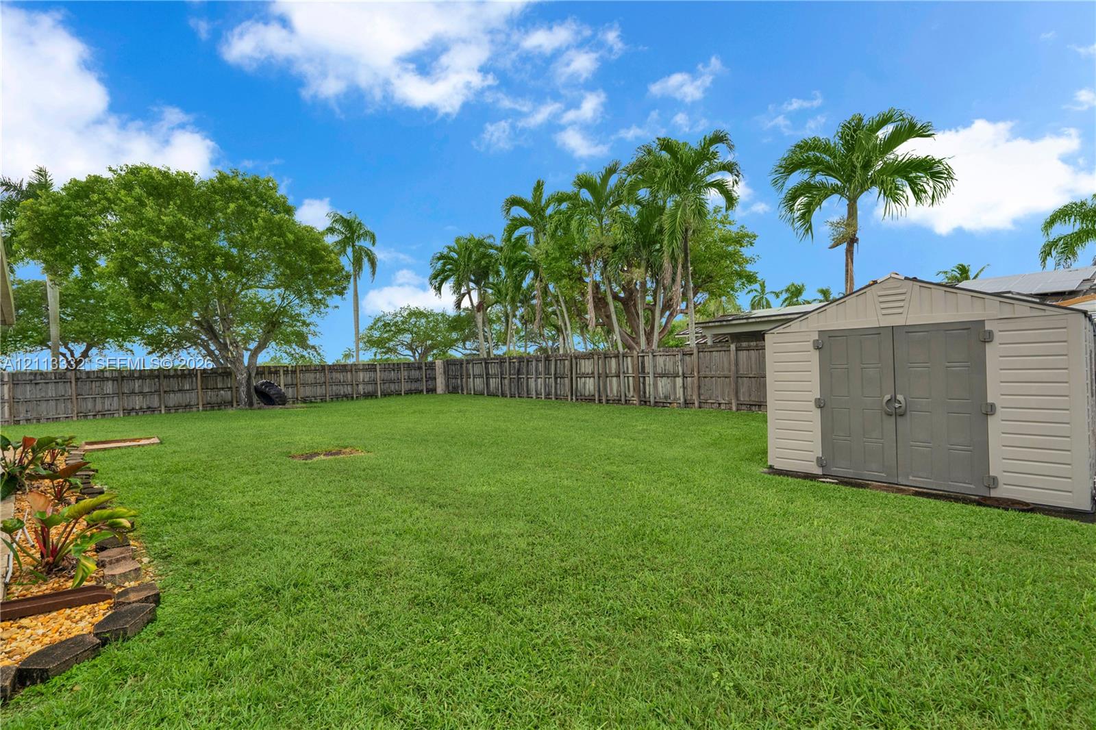 Expansive backyard featuring a manicured lawn, full-perimeter wood fencing, mature palm trees, and an exterior storage shed