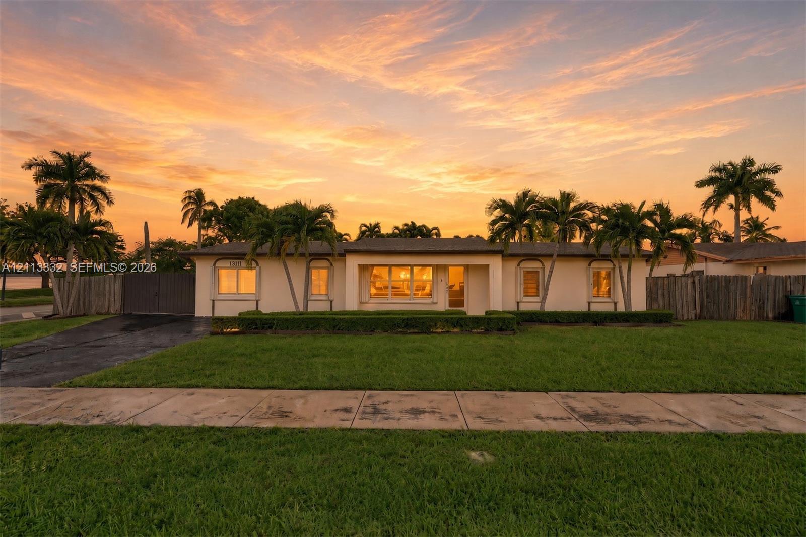 Single-story residence featuring a stucco exterior, expansive lawn, mature palm trees, manicured hedges, and a paved driveway