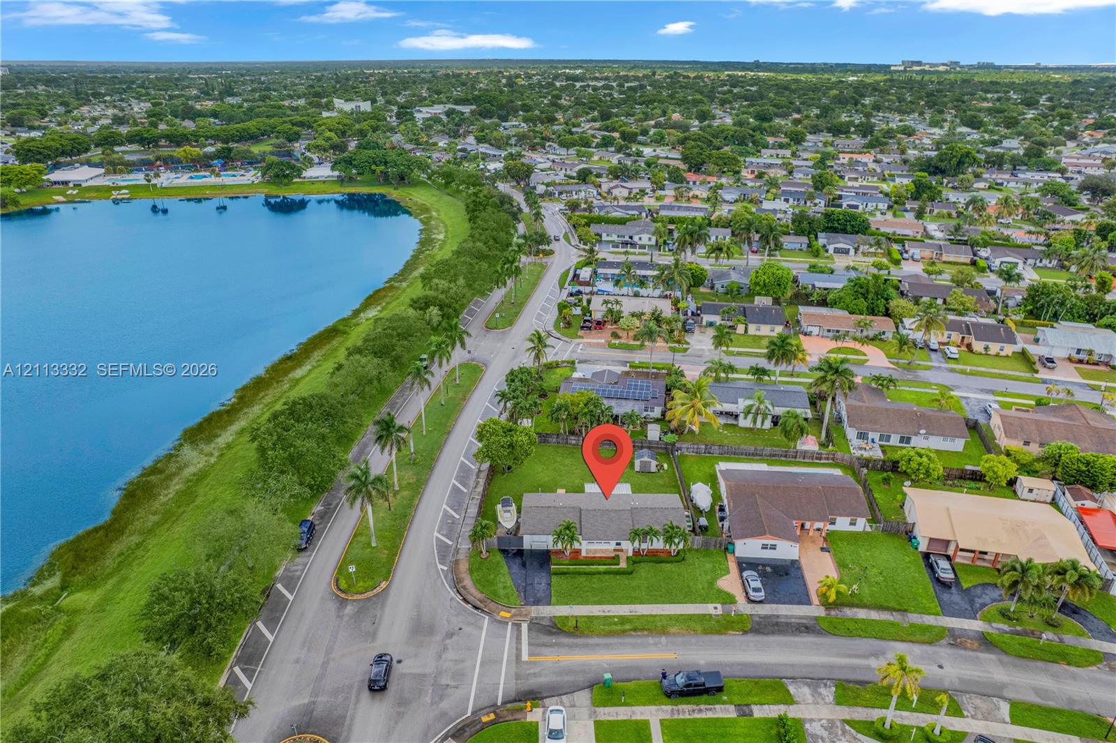 Aerial perspective featuring a single-story residence situated on a lot bordering a large lake with expansive lawn space and mature palm trees